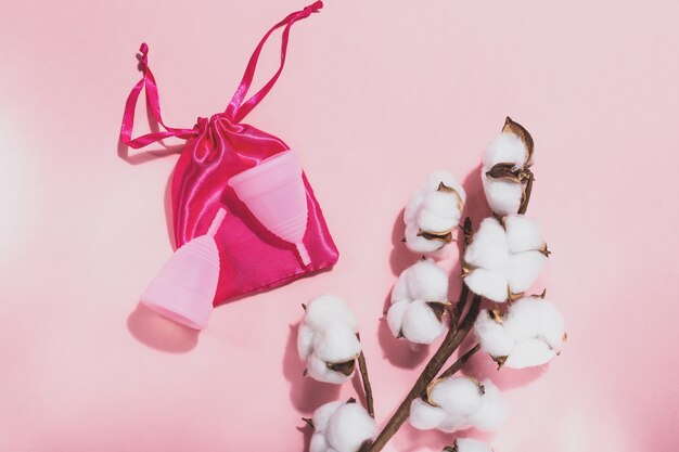 Two pink menstrual cups on a pink cloth bag and a branch of cotton on a pink background