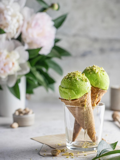 Two pistachio ice cream cones in a glass with chopped pistachios and flowers in the back