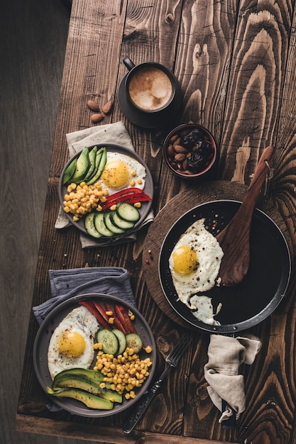 Two plates and a frying pan with cooked eggs with avocado, paprika, cucumber and canned corn on a dark wooden background. Healthy breakfast. Top view with copyspace. Flat lay.