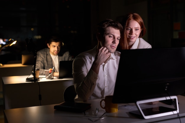 Two pleasant business people working together on computer, discussing project in office at late night. side view on redhead lady and handsome guy colleagues brainsotrming. in dark boardroom