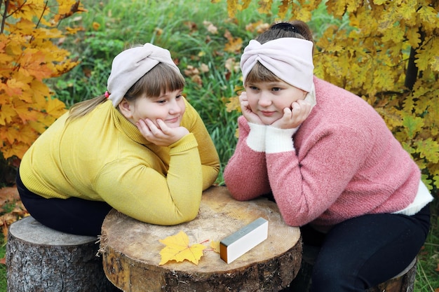 Two plump smiling girls sit on stumps outdoors in autumn and listen to music