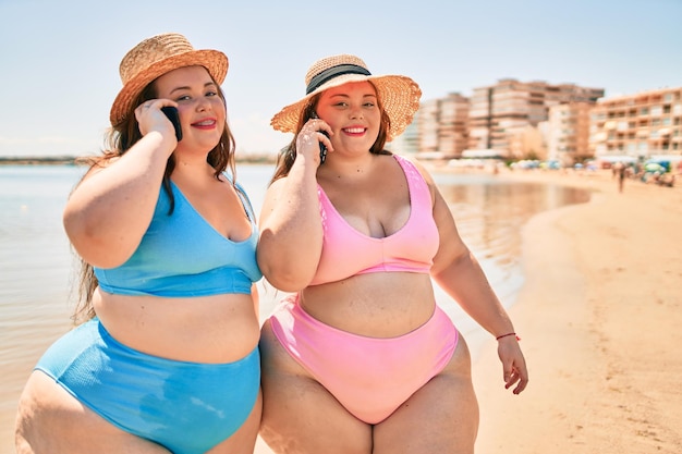 Two plus size overweight sisters twins women speaking on the phone at the beach on summer holidays