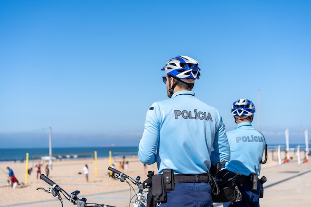 Two policeman patrolling seaside promenade on bicycles People are sunburning on the city public beach on the Atlantic shore