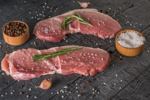Два стейка из свинины с мисками перца и соли на деревянном столе. Ингредиенты для приготовления мясных блюд.