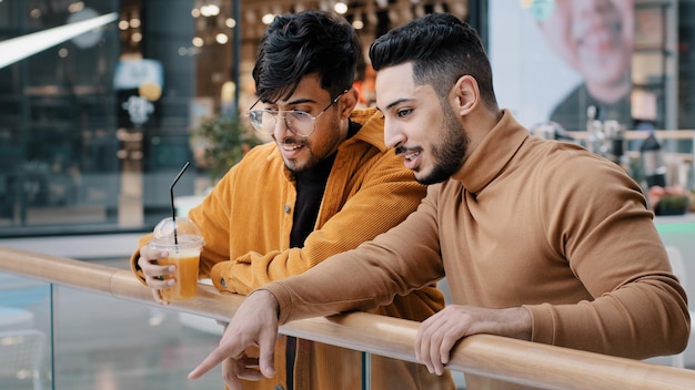 Two positive arab guys friends stand in public place in shopping center communicate pointing fingers