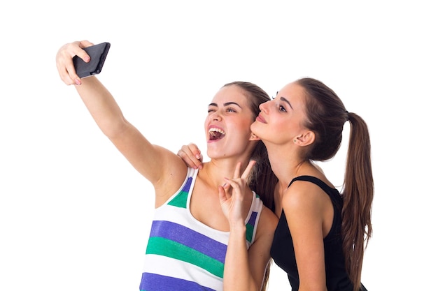 Two positive young women in striped and black shirts makin selfie on white background in studio