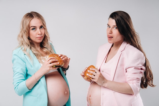 Two pregnant women in suits with hamburgers in their hands on a gray background.