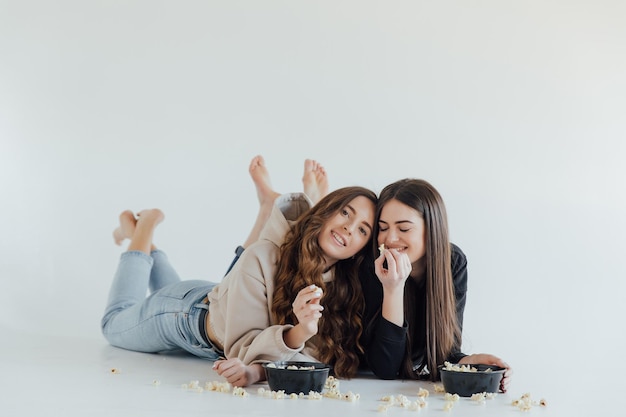 Two pretty cheerful young girls friends standing isolated over white background, eating popcorn