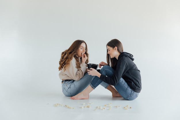 Two pretty cheerful young girls friends standing isolated over white background, eating popcorn