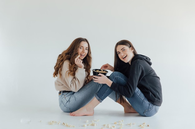 Two pretty cheerful young girls friends standing isolated over white background, eating popcorn