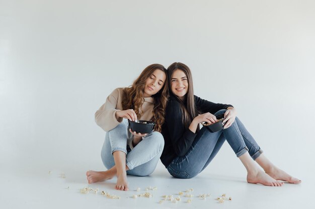 Two pretty cheerful young girls friends standing isolated over white background, eating popcorn