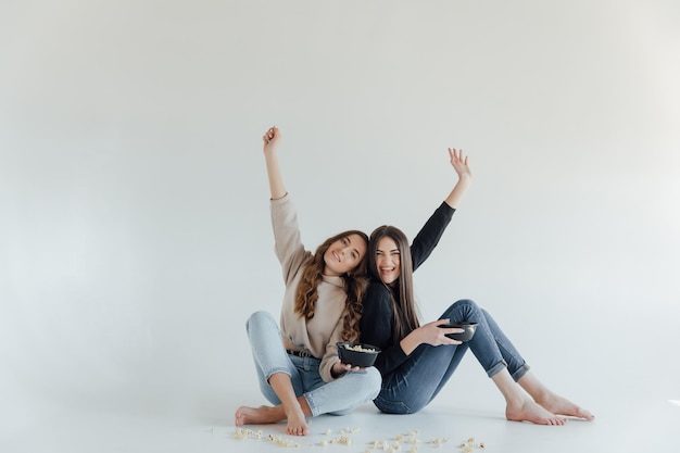 Two pretty cheerful young girls friends standing isolated over white background, eating popcorn