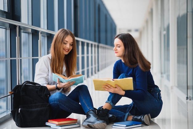 Two pretty female students with books sitting on the floor in the university hallway