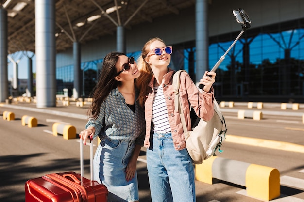 Two pretty girls in sunglasses happily taking photo on cellphone together with suitcase and backpack on shoulder outdoor near airport