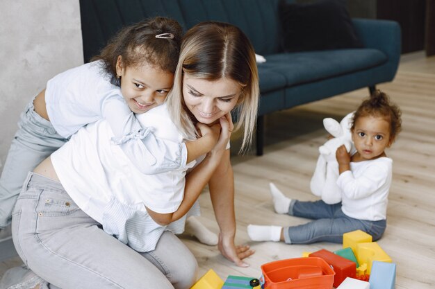 Two pretty kids and their blonde mother sit on floor and play with toys near sofa Afroamerican sisters playing at home