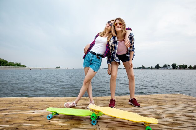 市松模様のシャツとデニムのショートパンツを着た2人のかわいい笑顔のブロンドの女の子が桟橋に立っています