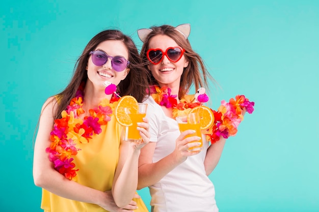 Two pretty smiling caucasian girls wears tshirts with lei and sunglasses hold orange cocktails on blue background