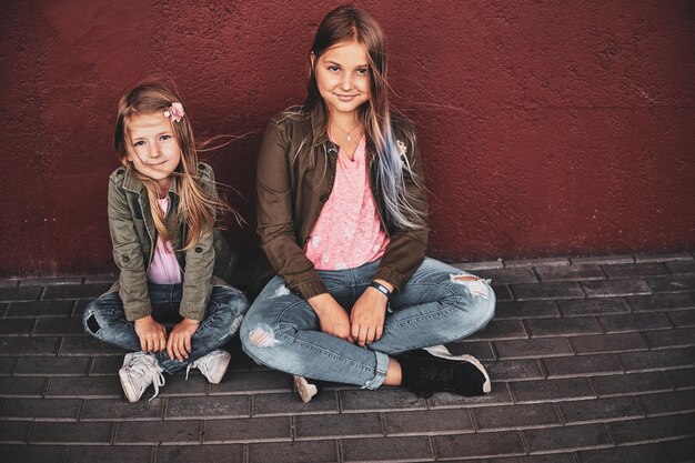 Due belle ragazze sorridenti sono sedute sul pavimento vicino al muro rosso sulla strada.