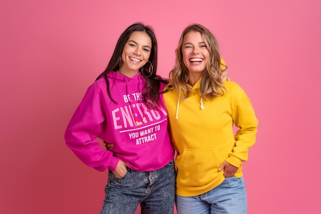 Two pretty women friends embracing having fun together, blond and brunette, wearing hoodie posing on pink isolated