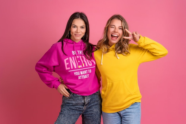 Two pretty women friends embracing having fun together, blond and brunette, wearing hoodie posing on pink