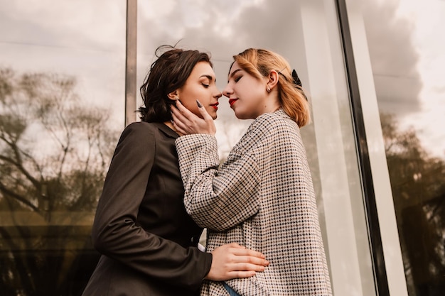 Two pretty women friends posing near glass building. Couple of gay lesbian girls hugging embracing