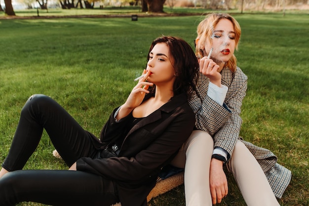 Two pretty women friends sitting on grass smoking cigarette. Couple of gay lesbian girls hugging