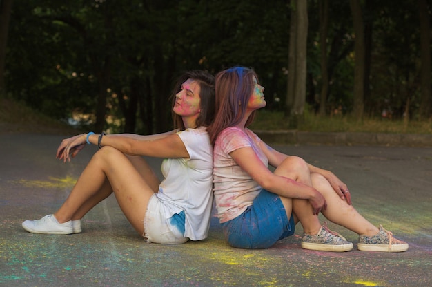 Two pretty young friends sitting on the asphalt covered with powder Holi