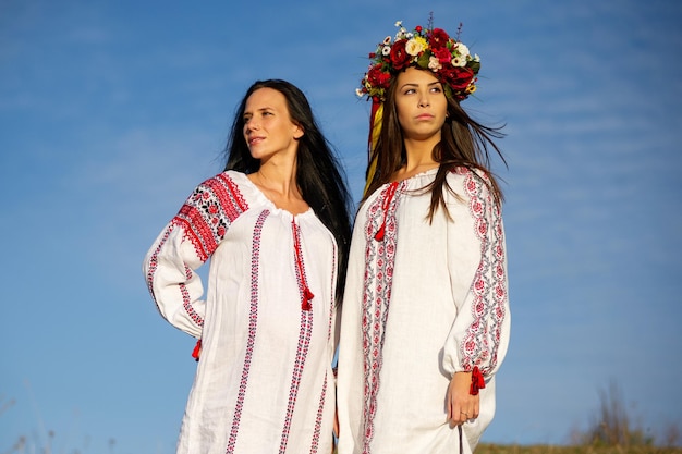두 명의 예쁜 젊은 여성이 전통적인 우크라이나 옷을 입고 꽃 화환을 입고 밀밭을 걷고 있습니다. 손으로 장식된 꽃 왕관을 쓴 아름다운 민족 소녀는 자연을 감상합니다.