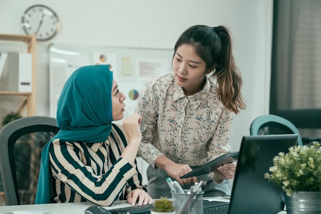 two pretty young multi ethnic businesswomen discussing over digital tablet in modern office. muslim female manager explain project of work to colleague in studio. creative team work partners concept.