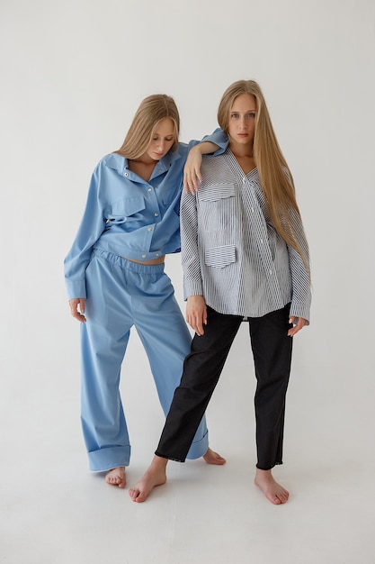 Two pretty young twin sisters with long blond hair posing on white background in oversize clothes. Fashion photoshoot