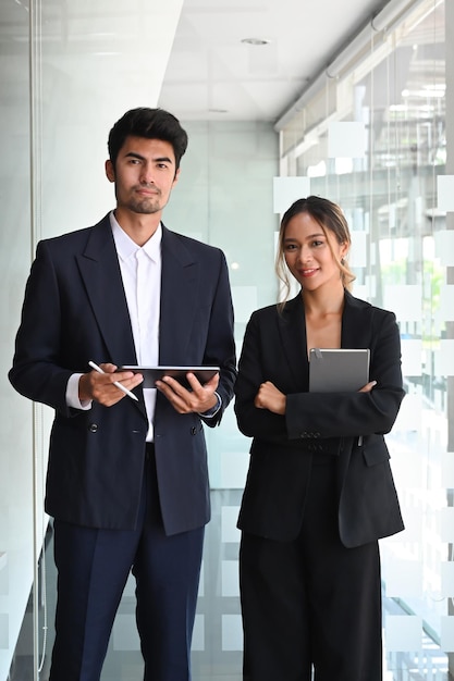 Two professional businesspeople holding digital tablet and looking at camera while standing in office lobby hall