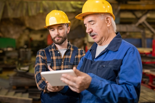 Two professional engineers in helmets and workwear looking at tablet display while discussing technical data