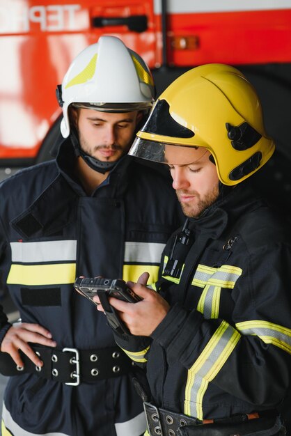 Two professional firefighters with uniforms and protective helmets posing infront of a firetruck