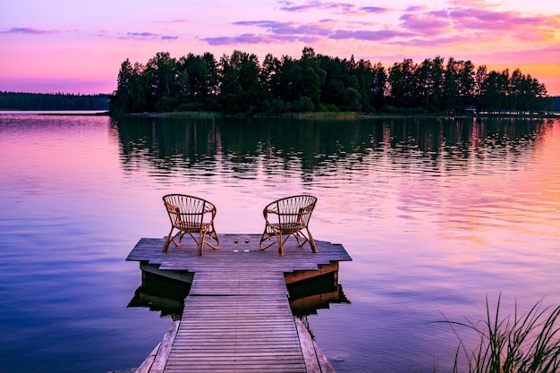 Photo two rattan wooden chairs and glasses of red wine on a pier overlooking a lake at sunset in finland