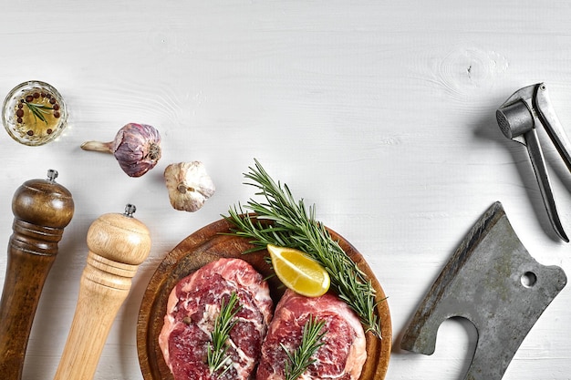 Two raw beef steak with spices on wooden board and kitchen ax on white background top view