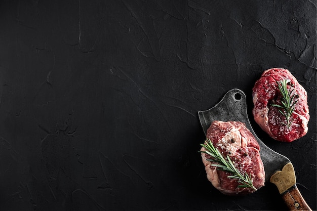 Two raw fresh ribeye steaks with salt pepper rosemary garlic and an ax on black background