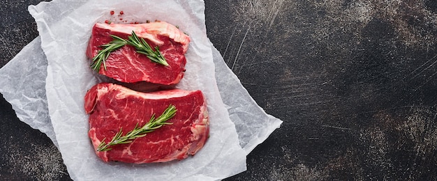 Two raw steak New York  with rosemary and spices on a piece of parchment paper on old dark stone background. Top view. Mock up.