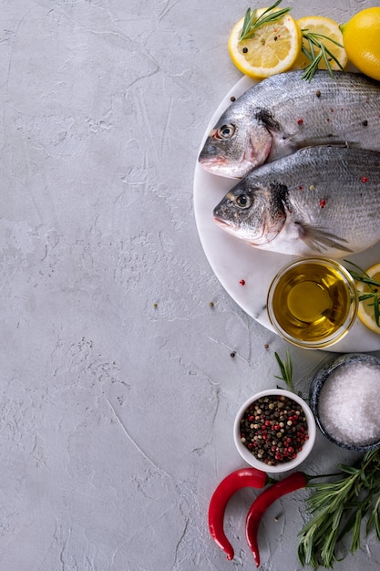 Two ready to cook raw bream fish with herbs, lemon and olive oil on marble board on gray background. With copy space. Top view