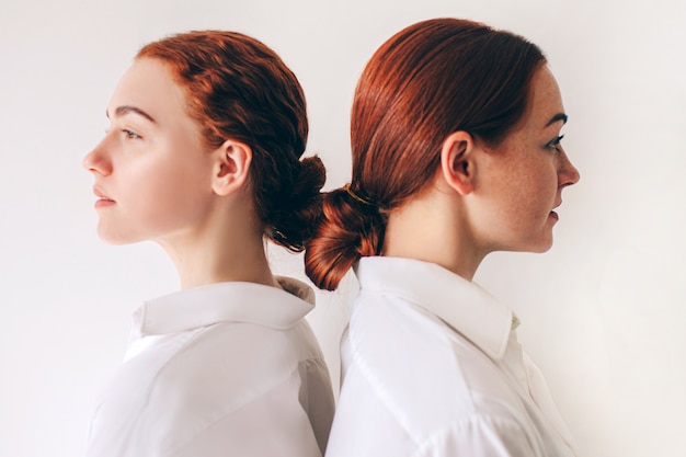 Two red-haired sisters stand isolated on a white background in shirts. Women standing with their backs to each other. hair is twisted into a bundle. They have straight hair and curly curls.