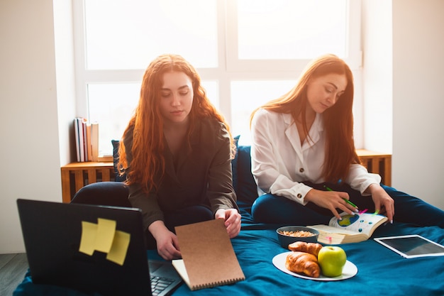 Two red-haired students study at home or prepare for exams. Young women doing homework in a dormitory bed near the window. There are notebooks, food books, a tablet and laptop and documents