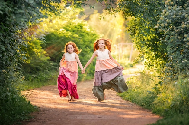 Two redhaired girls in long dresses run barefoot along the path in the park towards the sunset view from the back