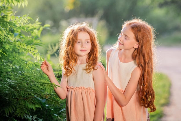 Two redhead sisters in long linen dresses are walking in park on sunny summer day