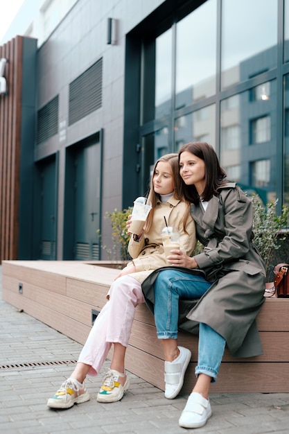 Две спокойные женщины в элегантной повседневной одежде пьют, сидя у современного здания и наслаждаются отдыхом