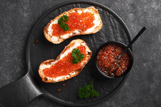 Two sandwiches with red caviar Salmon red caviar in bowl and sandwiches server on old iron plate on old black table background Top view Copy space