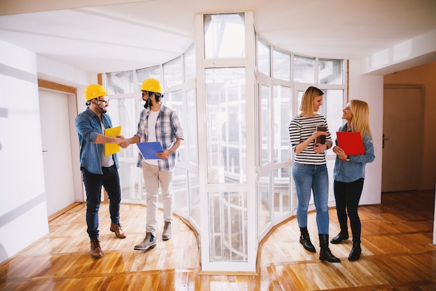 Two satisfied young professional engineers with helmets shaking hands after making a deal about the project while on the right side two smiling business women holding folders and talking.