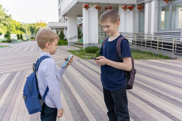 Two schoolboys with backpacks stand in background of the school and chatting on the phone to each