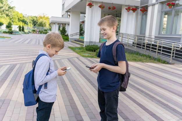 Two schoolboys with backpacks stand in background of the school and chatting on the phone to each