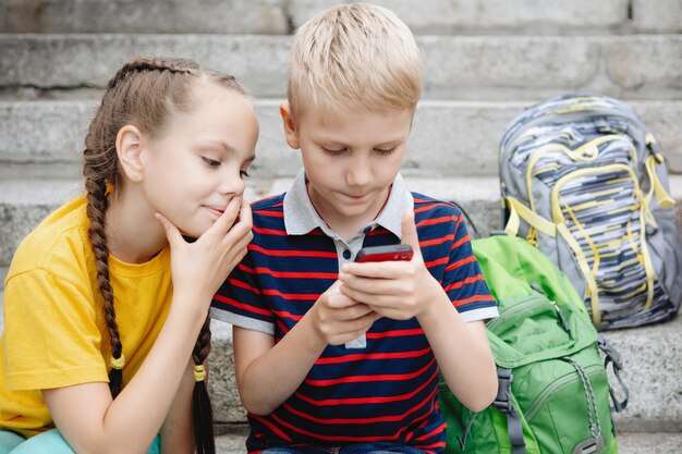 Two schoolchildren, boy and girl are sitting on the steps and looking at the smartphone.