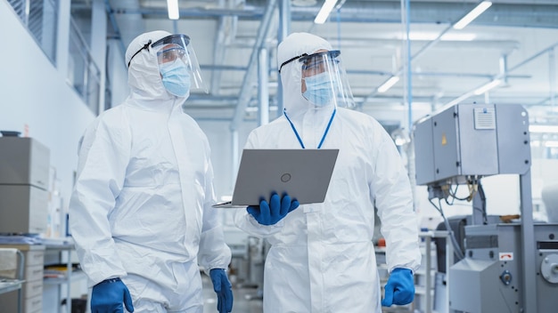 사진 두 명의 과학자가 무거운 산업 공장에서 살균 커버와 얼굴 마스크를 입고 노트북 컴퓨터를 사용하여 산업 기계 설정을 조사하고 생산 기능을 구성합니다.