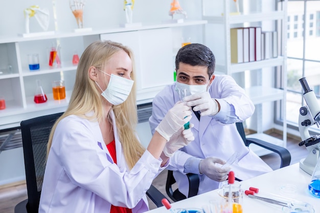 Two scientists wears hygiene protective mask are working holding looking at test tube with sample in a chemistry lab scientist are doing investigations in Laboratory analysis background